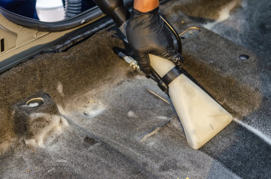A professional cleaner wearing a black glove is using a handheld extraction tool to deep clean a residential carpet in a living room. The carpet appears to be a dark-colored, textured surface with visible soap suds and foam from the cleaning process. The extractor nozzle is fitted with a white, flexible hose connected to a cleaning machine, partially visible at the top of the image. The surrounding area shows a corner of the room with a light wooden baseboard and various electrical cables running along the edge of the carpet. The room is well-lit, highlighting the wet and cleaned area of the carpet as part of a surface cleaning or sanitisation procedure provided by Carpet Cleaners SW5, suitable for domestic cleaning and maintaining hygiene in the home.