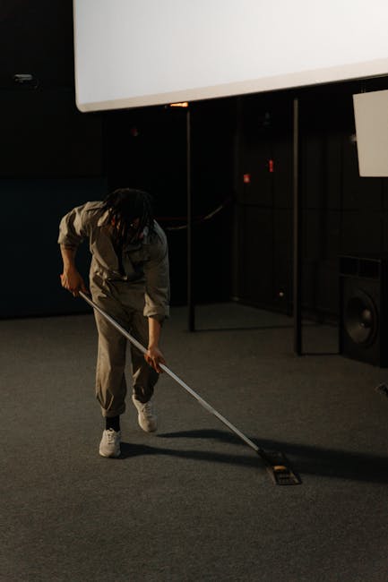 A professional cleaner wearing casual work attire is performing surface cleaning on a carpeted floor in a dark indoor setting, possibly a conference room or cinema space. The cleaner is using a long-handled tool to vacuum or shampoo the grey carpet, which appears neat and well-maintained. In the background, there is a large white projection screen mounted on the wall, and a black speaker is visible on the side, indicating a space used for presentations or events. The lighting is dim, highlighting the cleanliness and the focus on the detailed carpet cleaning process. Carpet Cleaners SW5 specializes in deep cleaning and sanitisation services for commercial spaces in the Earls Court SW5 area.