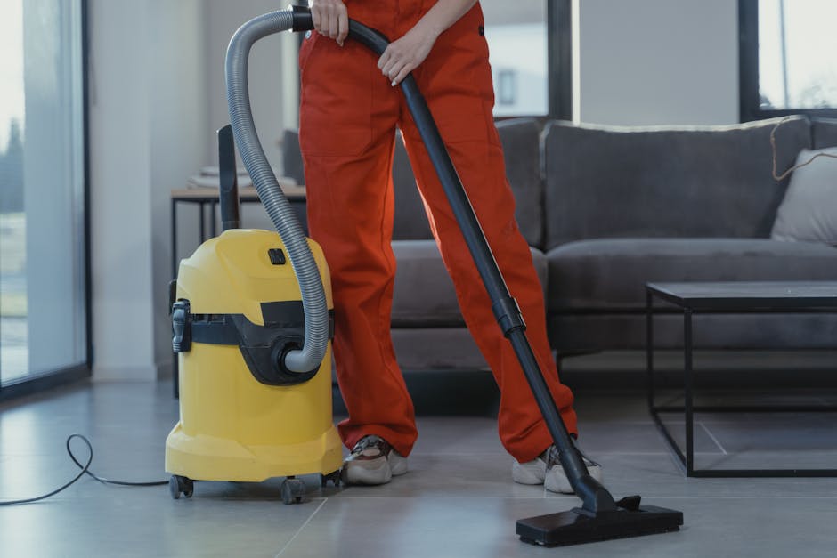 A professional cleaner wearing a black glove is using a handheld extraction tool to deep clean a residential carpet in a living room. The carpet appears to be a dark-colored, textured surface with visible soap suds and foam from the cleaning process. The extractor nozzle is fitted with a white, flexible hose connected to a cleaning machine, partially visible at the top of the image. The surrounding area shows a corner of the room with a light wooden baseboard and various electrical cables running along the edge of the carpet. The room is well-lit, highlighting the wet and cleaned area of the carpet as part of a surface cleaning or sanitisation procedure provided by Carpet Cleaners SW5, suitable for domestic cleaning and maintaining hygiene in the home.