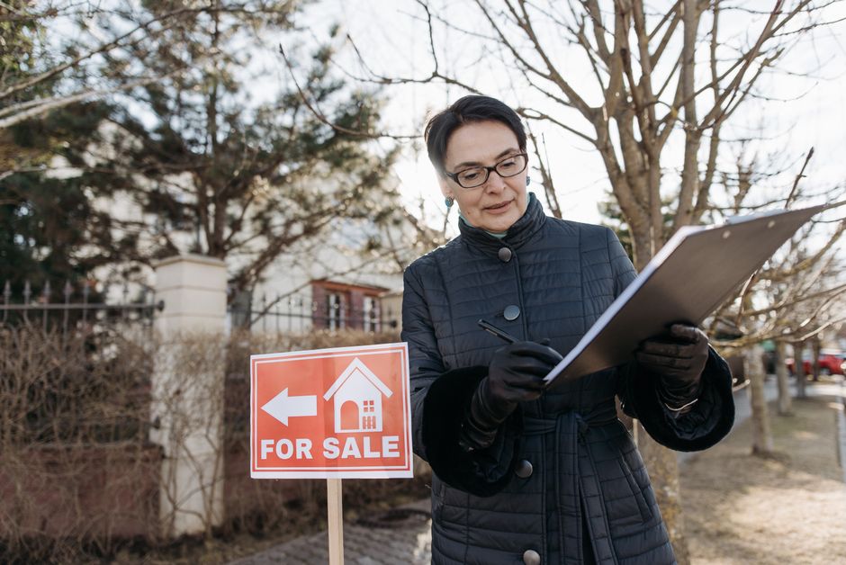 A professional cleaner in a black suit and glasses is presenting a detailed home inspection checklist printed on a clipboard to a man and a woman in an office setting. The room features bright natural